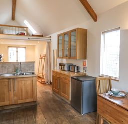 A kitchen with wooden cabinets a small refrigerator a dining table set with dishes and a loft area above at The Loft Cottage in Totnes
