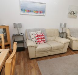 A living room with two beige sofas, cushions, side tables with lamps, a wooden dining table with chairs, and wall paintings at Badger Sett in Granite Henge near Looe
