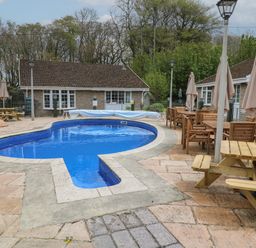 An outdoor pool area with wooden tables chairs umbrellas and cottages in the background at Badger Sett in Granite Henge near Looe