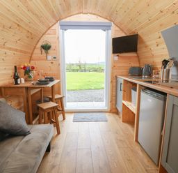 A small wooden kitchen and dining area with a sofa a table with stools kitchen appliances and an open door showing a grassy outside view at Nebo Pod in Amlwch