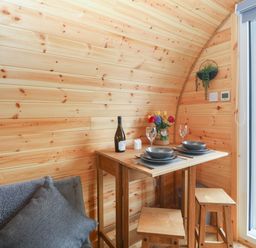 A small wooden dining table set with plates glasses and a bottle of wine next to a sofa and an open door at Nebo Pod in Amlwch
