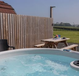 An outdoor area with a hot tub in the foreground a wooden picnic table with tableware and chairs next to a wooden fence at Nebo Pod in Amlwch