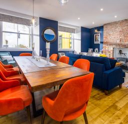 A dining area with orange chairs around a wooden table next to a living room with blue sofas and a brick fireplace at The Indigo House in Berwick-Upon-Tweed