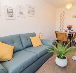 A blue sofa with yellow cushions a wooden table with a potted plant and a dining table with brown chairs in a living room at Pebble Bay in Appledore