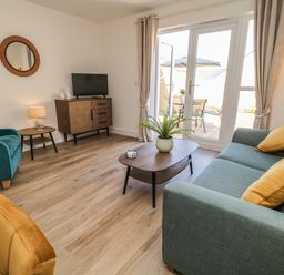 A living room with a teal sofa and yellow cushions a green armchair a wooden coffee table with a plant a cabinet with a television and patio doors with curtains at Pebble Bay in Appledore
