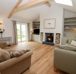 A living room with two sofas a fireplace wood logs shelves and a glass door at Cotswold View in Shipton Under Wychwood