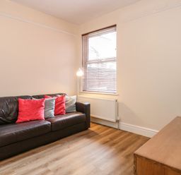 A living room with a brown leather sofa with red and gray pillows a floor lamp a window with blinds a radiator a wooden floor and a wooden TV stand with a television on it at Seaside House in Gorleston-On-Sea