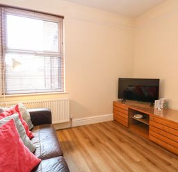 A living room with a brown leather sofa with red and gray cushions a wooden TV stand with a television and a window with wooden blinds at Seaside House in Gorleston-On-Sea