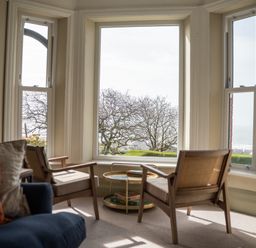 A sitting area with two wooden armchairs and a round side table near large windows overlooking trees and a distant view at Haulfryn in Barmouth