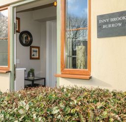 An open window and door with a view into a room with a small table and plant a sign on the wall saying Inny Brook Burrow at Inny Brook Burrow in Davidstow near Camelford