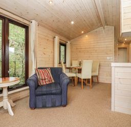 A living area with a blue armchair and a round wooden table next to large windows and a dining table with cream chairs in a wooden cabin Canadian Cabin No 39 Keswick