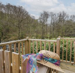 A wooden deck with a straw hat on the railing overlooking trees and a river at Pod No 6 Keswick