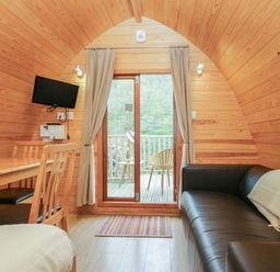 A wooden pod interior with a black sofa beside a table with chairs and a door leading to a balcony with chairs at Pod No 28 in Keswick