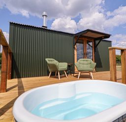 A wooden deck with green chairs and a small table outside a green corrugated metal building with a hot tub in the foreground at The Coop in Aston near Woore