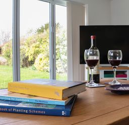 A wooden table with three books a bottle of wine two glasses of wine and a plate in a room with sliding glass doors at Little Chapter in Walberswick