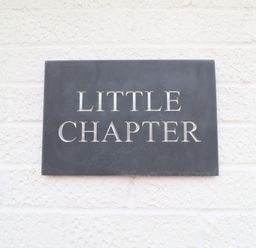 A black rectangular sign with the words Little Chapter mounted on a white textured wall at Little Chapter in Walberswick