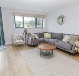 A living room with a grey sectional sofa pillows and blankets a round wooden coffee table an armchair a floor lamp a rug a round wall mirror and a window at Sebastian's View in Falmouth