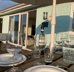 An outdoor dining table set with plates glasses a bottle of wine and a water jug on a wooden deck outside a building at Little Beach in Seaview