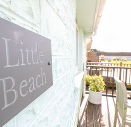 A sign reading Little Beach mounted on a light textured wall next to a wooden deck with a potted plant and outdoor furniture at Little Beach in Seaview