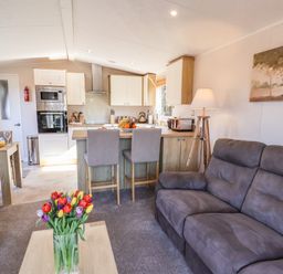 A living room with gray sofas and a wooden coffee table with flowers next to a kitchen with a breakfast bar and stools at Marigold Lodge in Runswick Bay near Staithes