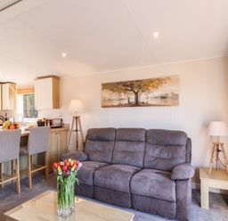 A living room with a brown sofa a coffee table with flowers two lamps stools at a kitchen counter and a painting on the wall at Marigold Lodge in Runswick Bay near Staithes