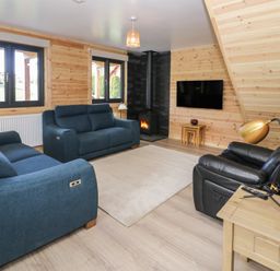 A living room with blue sofas a black leather chair a wood burning stove and a wall mounted tv at Sunset Lodge 5 in Llanbedr