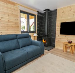 A living room with a blue sofa a wood burning stove a flat screen tv on the wall nested wooden tables and windows at Sunset Lodge 5 in Llanbedr