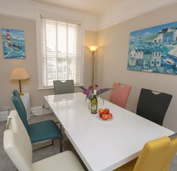 A dining room with a white table surrounded by six multicolored chairs and two paintings on the walls at Chine Heights in Bournemouth