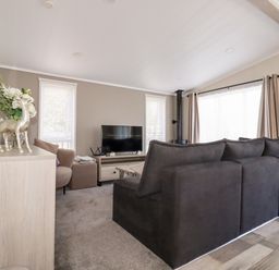 A living room with a large dark sofa a television two windows and a sideboard with decorations at 63 The Maples in Hewas Water near Sticker