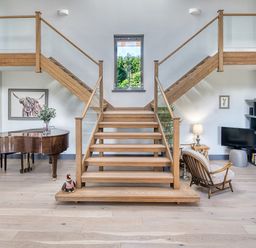 An interior with a wooden staircase leading up with glass railings a piano on the left and a reading area with a chair a lamp and a bookshelf on the right at Ben Luna Lodge Drumnadrochit Loch Ness