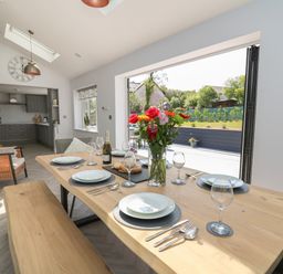 A dining area with a wooden table set for six with flowers and a bottle with an open plan kitchen in the background at Rhiwmynach in Pontarfynach near Aberystwyth