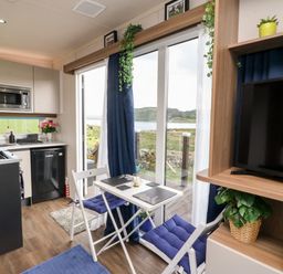 A small dining table with two chairs and blue cushions next to sliding glass doors in a kitchen with appliances and a television on a wooden stand with plants at Balvicar Beag in Balvicar Isle Of Seil
