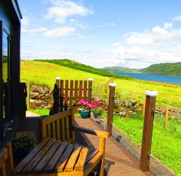 A wooden bench on a deck overlooking a grassy field and a body of water at Balvicar Beag in Balvicar Isle Of Seil