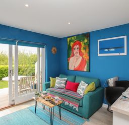 A living room with a blue sofa and colorful cushions and a small wooden table next to a glass door leading to a garden at Blue Waters in Weymouth