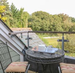 A small balcony with a round metal table and two chairs with cushions overlooking trees and a sports court at 5 Court Cottage Hillfield Village in Dartmouth
