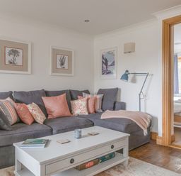 A living room with a grey sofa with pink and striped cushions and a white coffee table next to a bedroom at 5 Court Cottage Hillfield Village Dartmouth