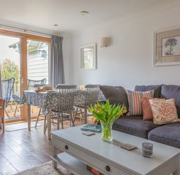 A living room with a gray sofa and a white coffee table with flowers and books and a dining table with chairs near glass doors leading to an outdoor area at 5 Court Cottage Hillfield Village in Dartmouth