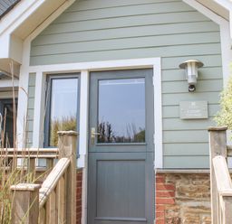 The front entrance of a cottage with a wooden door and side window with a wooden railing and bushes outside 5 Court Cottage Hillfield Village Dartmouth