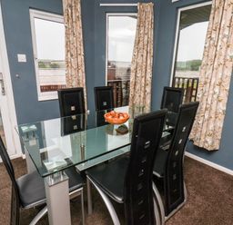 A dining room with a glass table surrounded by six black chairs and curtains on windows at Handale Banks Farm Lodge in Liverton