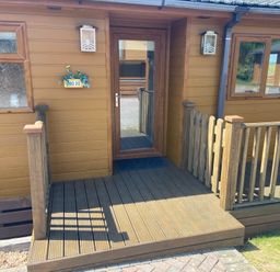 A wooden porch with a glass door and railings on a brown building at Mojo in Mullacott near Ilfracombe