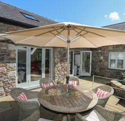 An outdoor patio area with a round wooden table and wicker chairs under a large umbrella at Traeth Ora in Moelfre