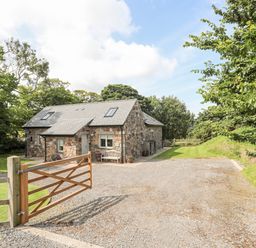 A stone cottage with a pitched roof surrounded by trees and a gravel driveway at Traeth Ora in Moelfre