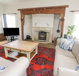 A living room with beige sofas and blue patterned cushions a wooden coffee table a red patterned rug a fireplace with a wood stove a television and windows at Wig Carna in Holyhead