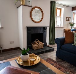 A living room with a black fireplace and a basket of firewood a blue sofa with pillows and a coffee table with a plant and candle at Fern Cottage Muddiford near West Down