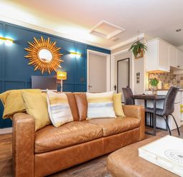 A living room with a brown leather sofa and yellow cushions next to a small dining area and white kitchen cabinets at Inny Brook Cottage in Davidstow near Camelford