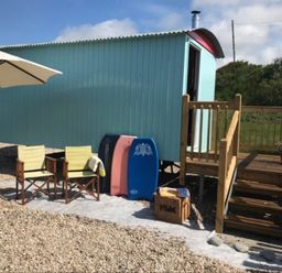 An outdoor area with a light blue wooden hut on stilts with stairs and railing two chairs under a large white umbrella and three bodyboards leaning against the hut at Shepherd's Hut in Southerndown near St Brides Major