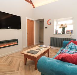 A living room with a teal sofa and red cushions a wooden coffee table a wall-mounted television and an electric fireplace at Mayfield Cottage Nr Malmesbury