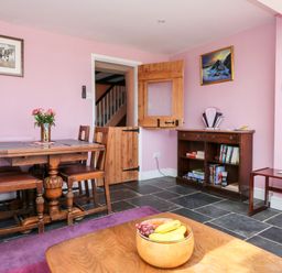 A dining area with a wooden table and chairs a wooden bowl with fruit on a coffee table and a wooden cabinet with books on shelves at The Old Forge Cottage in Kilkhampton