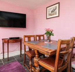 A dining area with wooden table and chairs a wall-mounted tv and a small wooden side table with a device at The Old Forge Cottage Kilkhampton