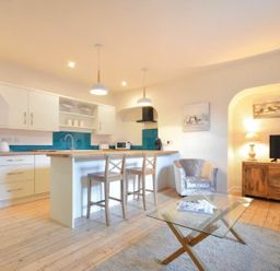 A kitchen area with white cabinets, a wooden countertop island with two wooden stools, a chair, a glass coffee table, and a wooden TV stand with a lamp and television at The Loft Amble